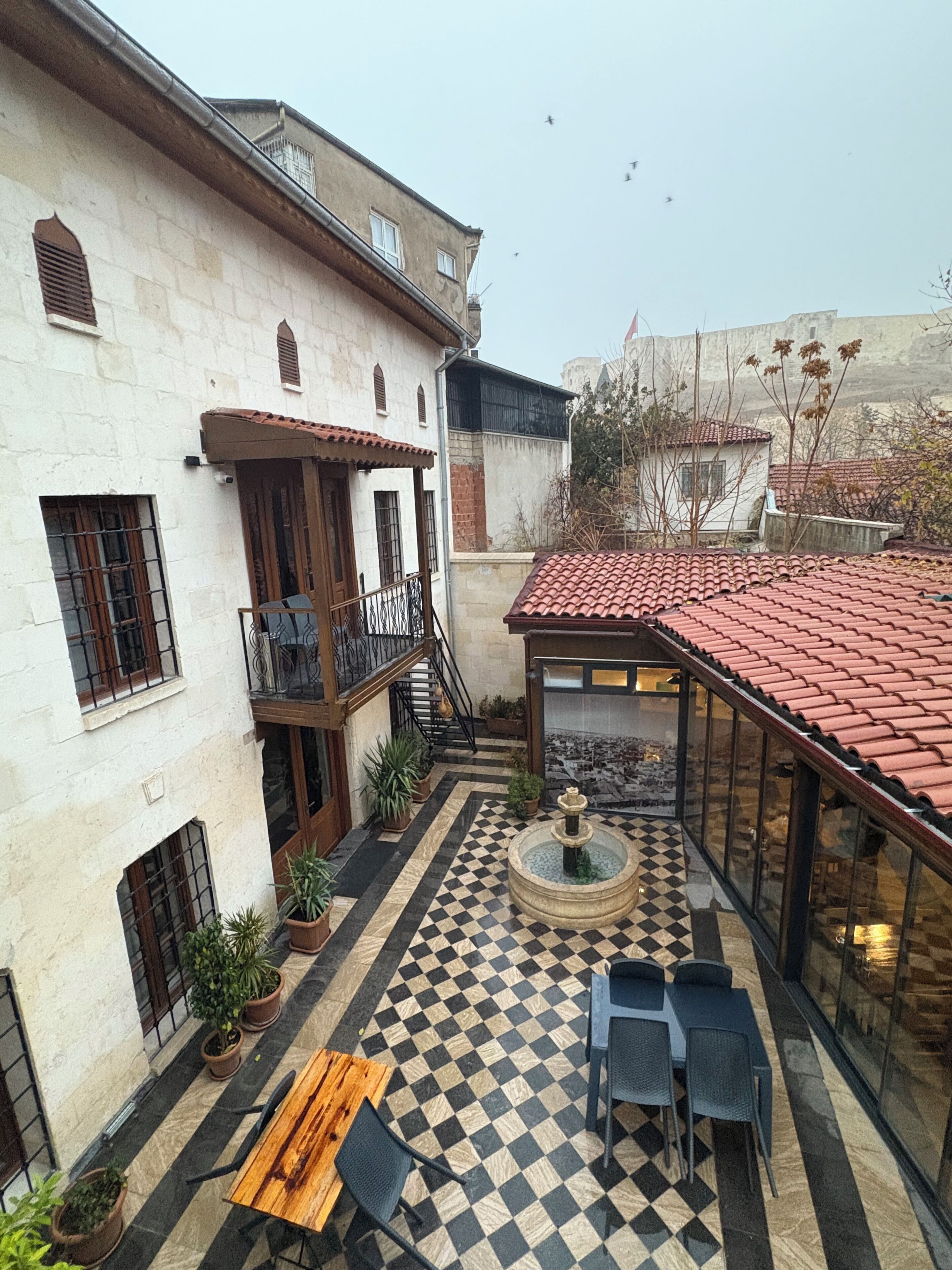 Un patio con suelo a cuadros, una fuente, mobiliario exterior y un edificio con balcón.
