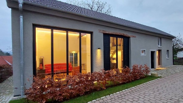 Une maison moderne avec de grandes fenêtres en verre, un canapé rouge à l’intérieur et un jardin bien entretenu.