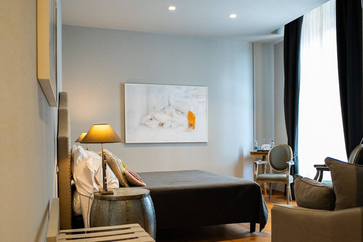 A modern hotel room with a bed, a desk, a chair, a lamp, and a painting on the wall.