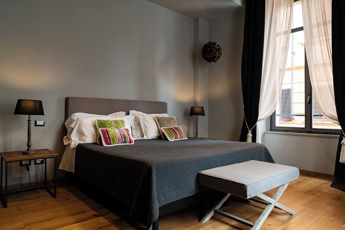 A modern bedroom with a large bed, two bedside tables with lamps, a bench, and a window with curtains.