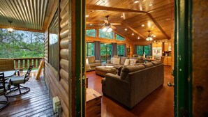 Una cabaña de madera con terraza, una sala de estar con sofá y comedor, y ventanas amplias con vistas al bosque circundante.