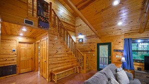 Interior de una cabaña de madera con una escalera, un sofá y un televisor.