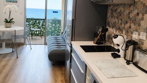 Ett modernt kök med utsikt över havet, en matplats med ett runt bord och stolar samt en grå soffa.