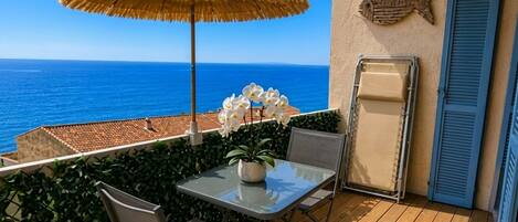 A balcony with a table, chairs, a potted plant, a straw umbrella, and a fish decoration on the wall.