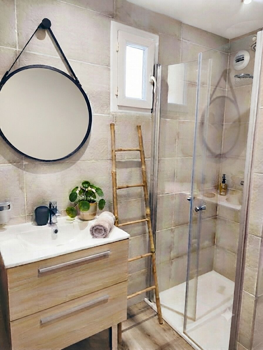 Une salle de bain avec un meuble-lavabo en bois, un miroir rond, une plante en pot, une serviette, une douche en verre et un escabeau.