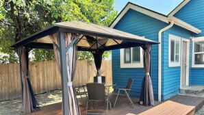 Eine überdachte Terrasse mit Tisch und Stühlen, ein Holzdeck und ein blaues Haus im Hintergrund.