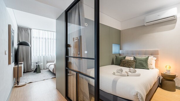 A modern hotel room with a glass partition, a bed with pillows, a bedside table with a lamp, and an air conditioning unit. - GuestReady - Exquisite stay in Matosinhos (Matosinhos)