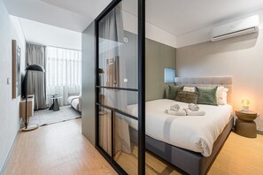 A modern hotel room with a glass partition, a bed with pillows, a bedside table with a lamp, and an air conditioning unit. - GuestReady - Exquisite stay in Matosinhos (Matosinhos)
