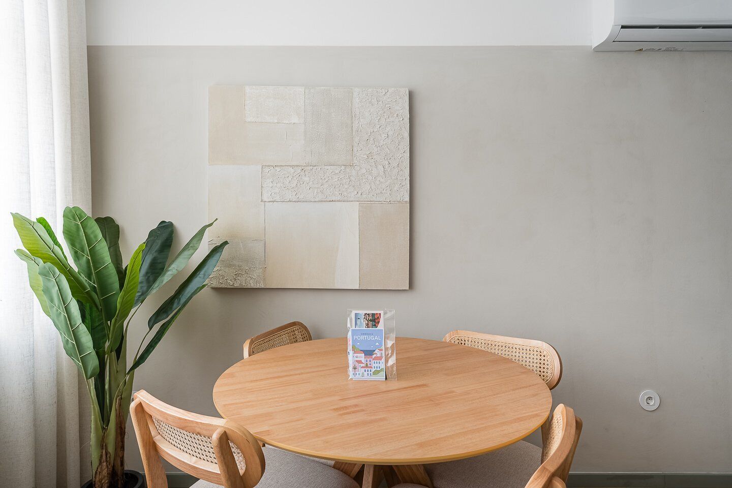 Una mesa redonda de madera con cuatro sillas de mimbre, una planta en maceta y una obra de arte abstracto enmarcada en la pared.
