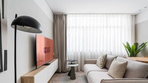 A modern hotel room with a flat-screen TV, a sofa, a small round table, and a large window with sheer curtains. - GuestReady - Exquisite stay in Matosinhos (Matosinhos)