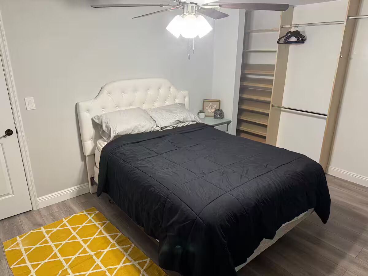 A bedroom with a tufted headboard, a black bedspread, a ceiling fan, a white wardrobe, and a yellow patterned rug.