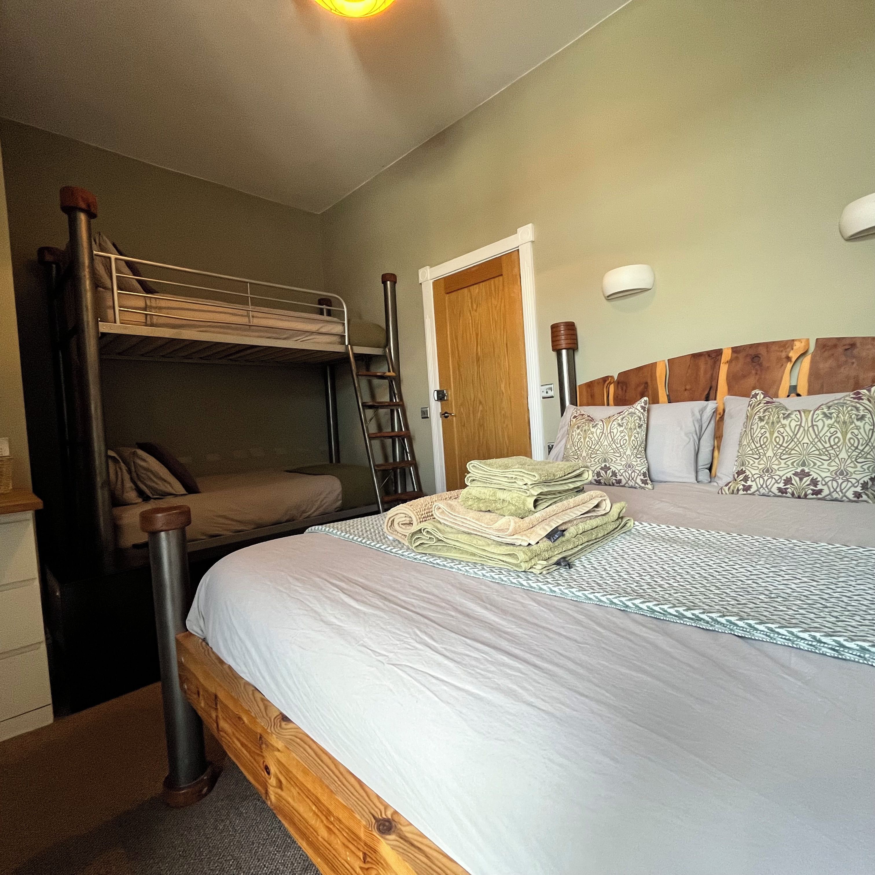 A bedroom with a bunk bed, a single bed, a wooden headboard, a bedside table, and a wall-mounted lamp.