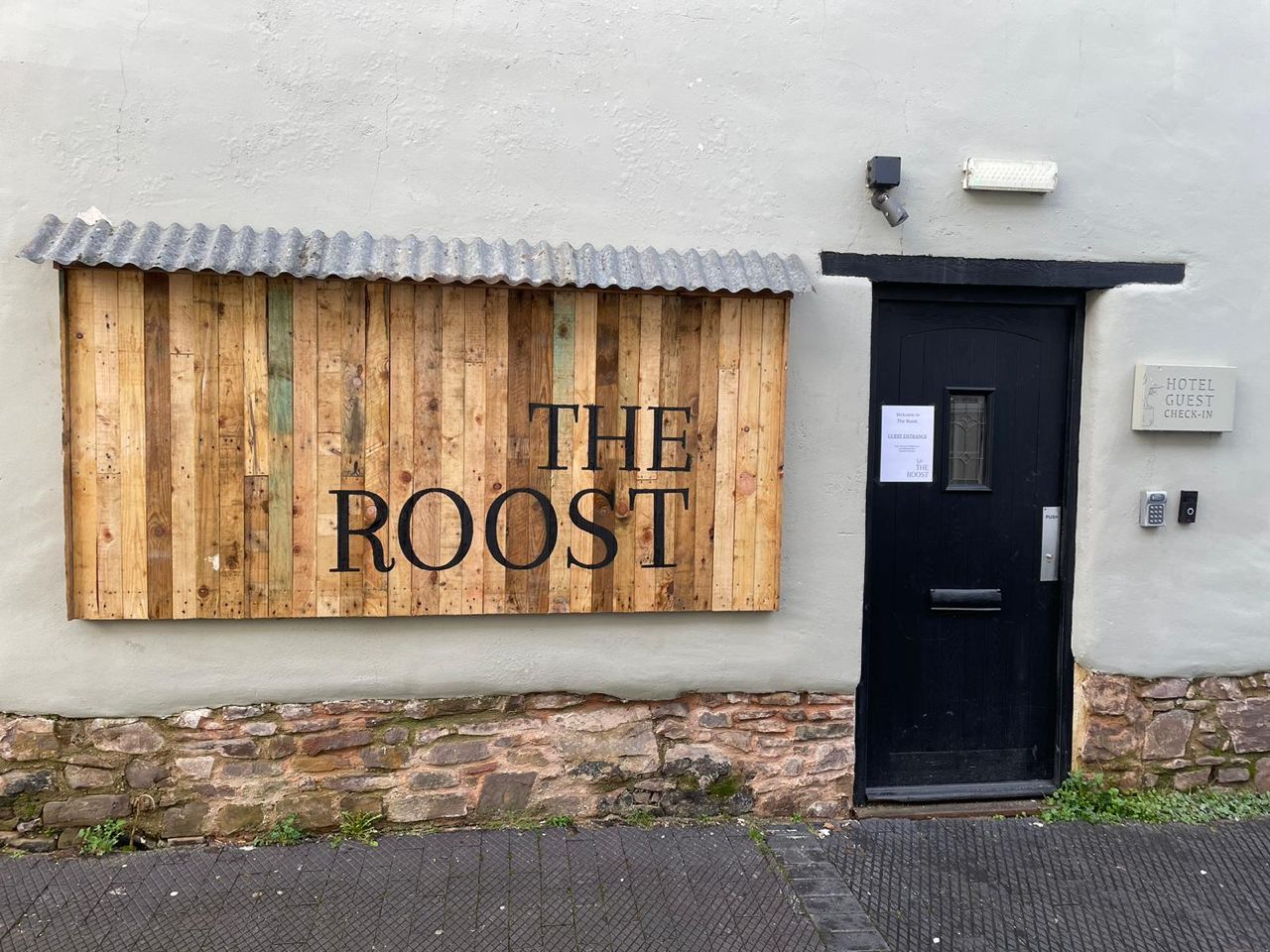 A building with a wooden sign reading 'THE ROOST' and a black door with a keypad lock.