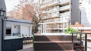 Un espace extérieur moderne aménagé avec un banc en bois et une table à plateau de verre, entouré d’une rambarde et offrant une vue sur un immeuble à plusieurs étages.