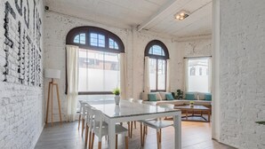 Une pièce spacieuse avec un mur en briques blanches, une table à manger avec des chaises, un canapé avec des coussins et de grandes fenêtres avec des rideaux.