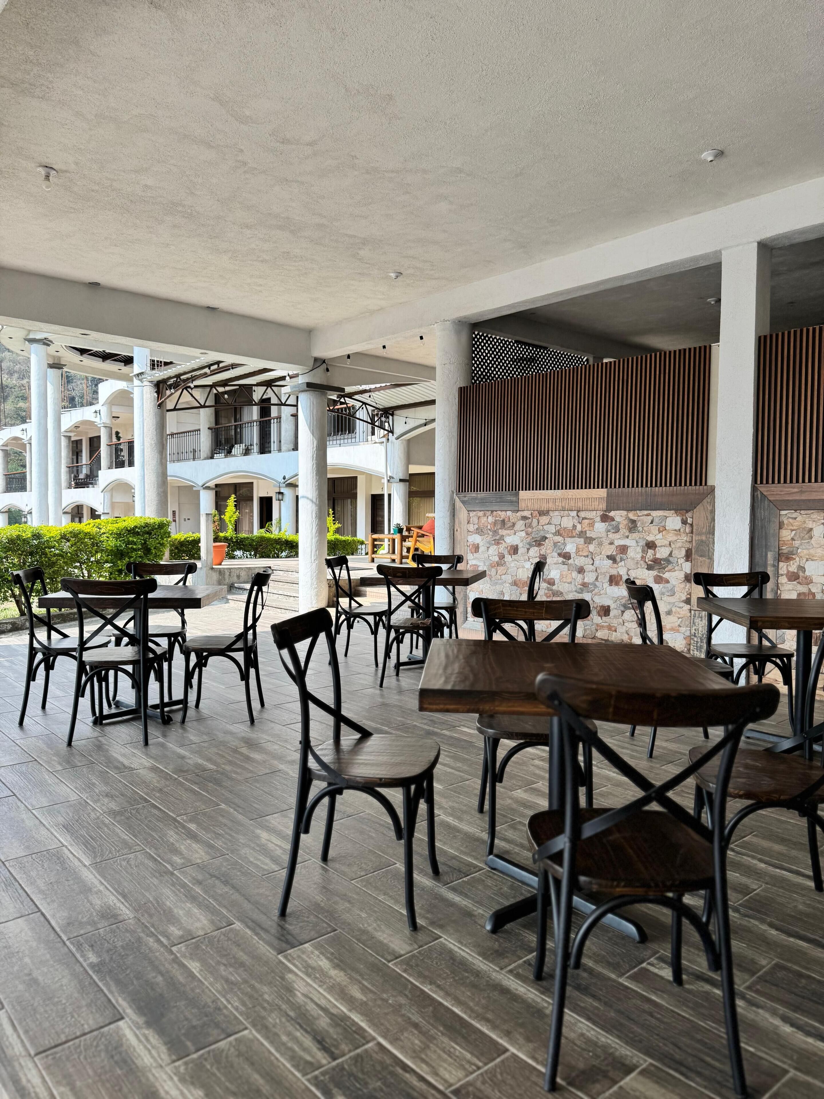 Un área de comedor al aire libre con varias mesas y sillas, un muro de piedra con diseño de listones de madera, y vista a un edificio con balcones al fondo.
