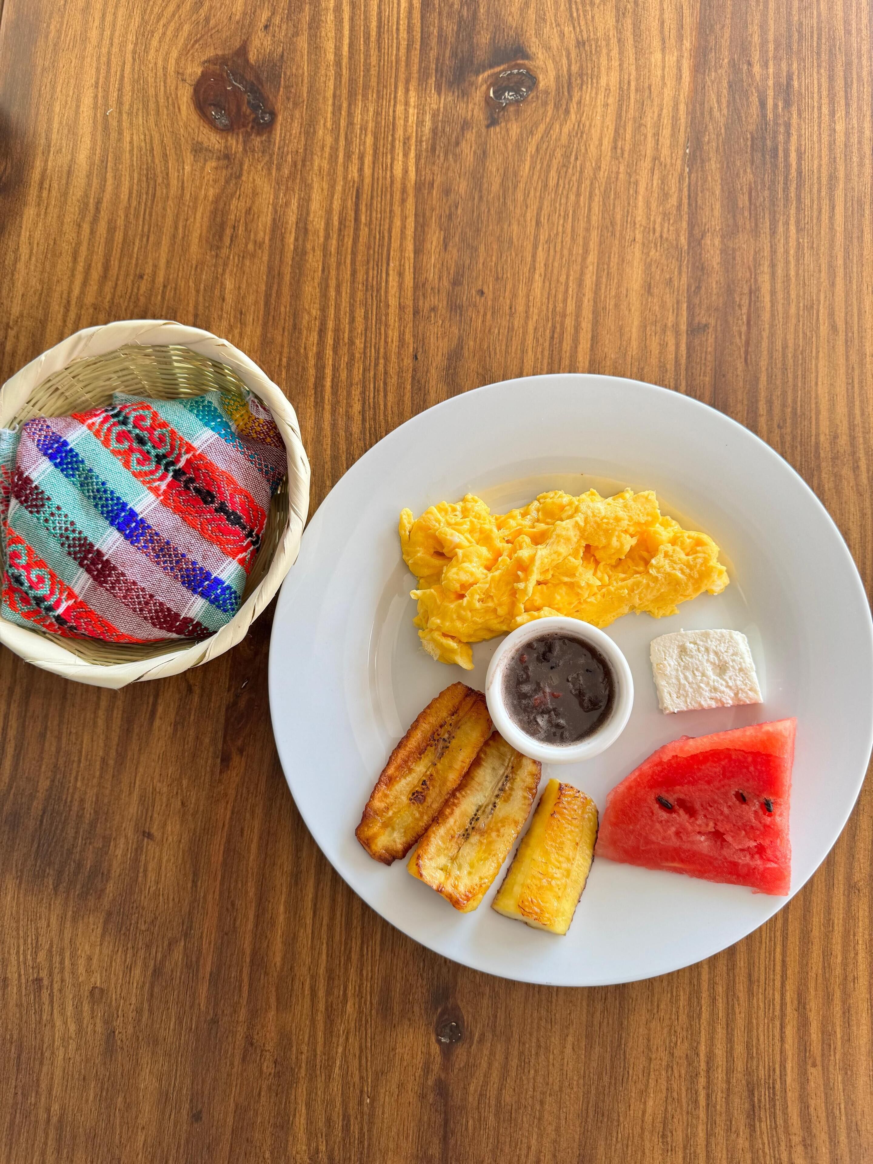 A plate with scrambled eggs, fried plantains, a slice of watermelon, and a small bowl of black condiment, alongside a woven basket with colorful fabric.
