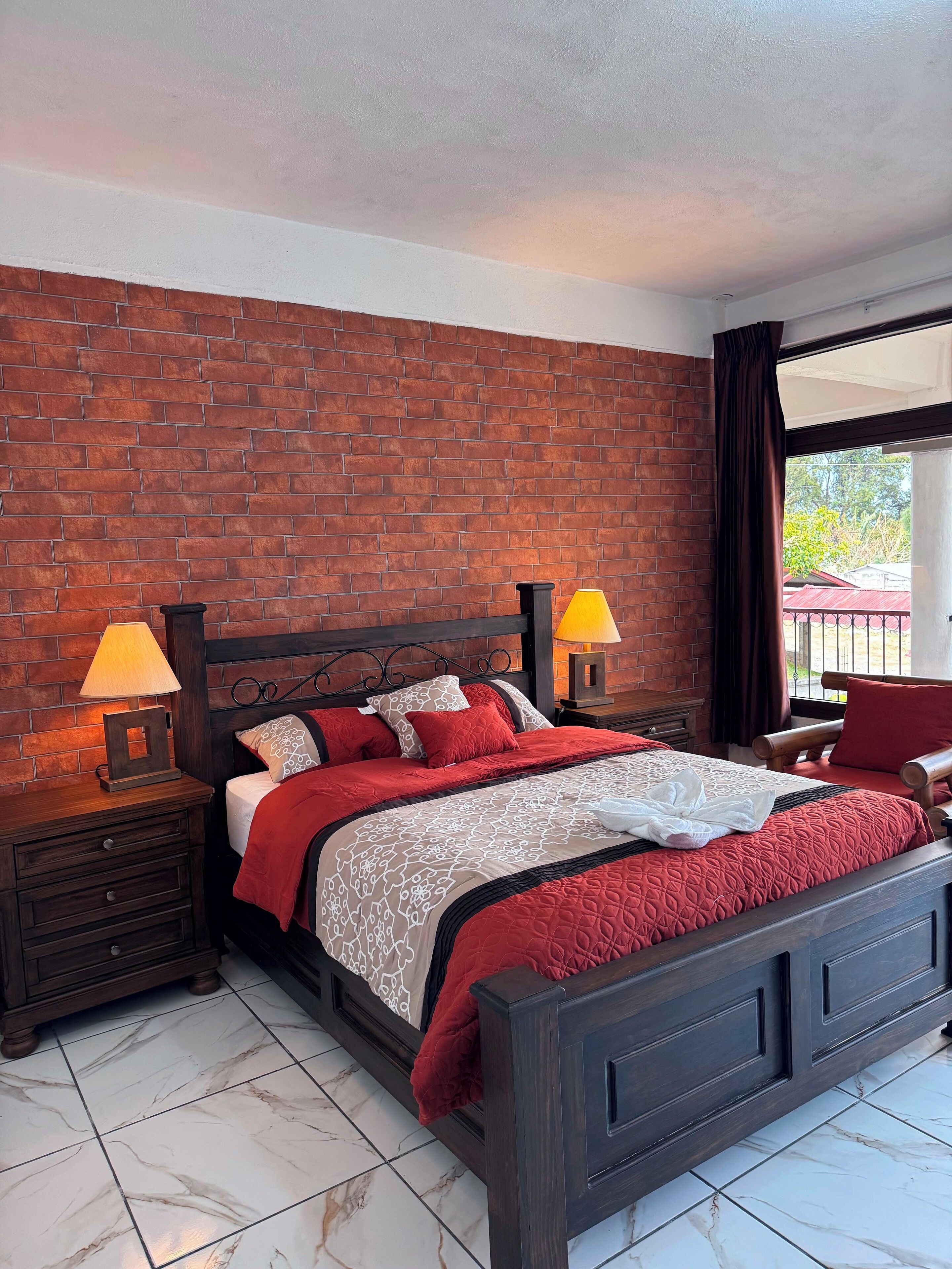 Un dormitorio con pared de ladrillo, cama con ropa de cama roja y blanca, dos lámparas de noche, una mesita de noche de madera y vistas al exterior a través de una ventana.