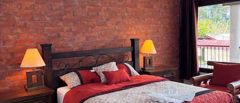 Un dormitorio con pared de ladrillo, cama con ropa de cama roja y blanca, dos lámparas de noche, una mesita de noche de madera y vistas al exterior a través de una ventana.