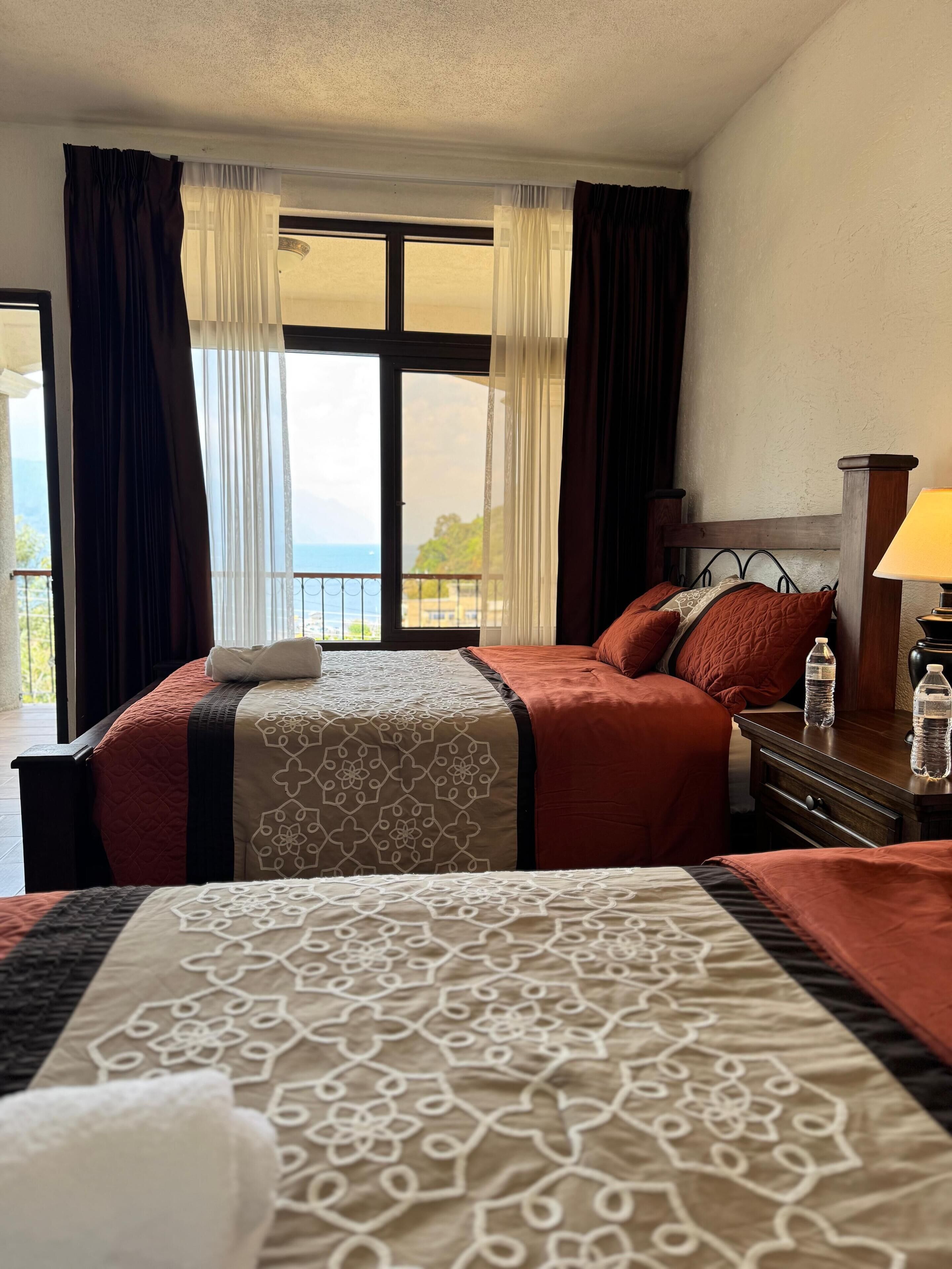 A hotel room with a large bed, a nightstand, a lamp, and a view of the sea through the window.