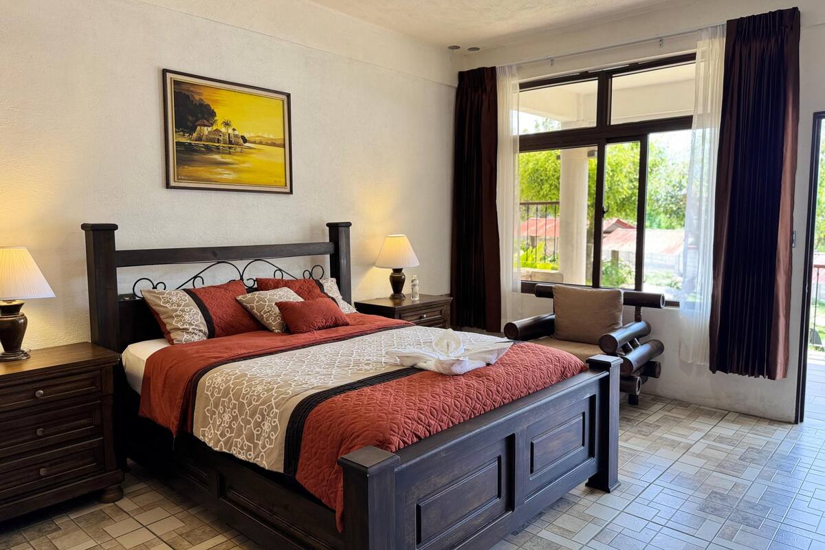 Een keurig opgemaakt slaapkamer met een bed, nachtkastjes, een stoel en een groot raam met gordijnen.