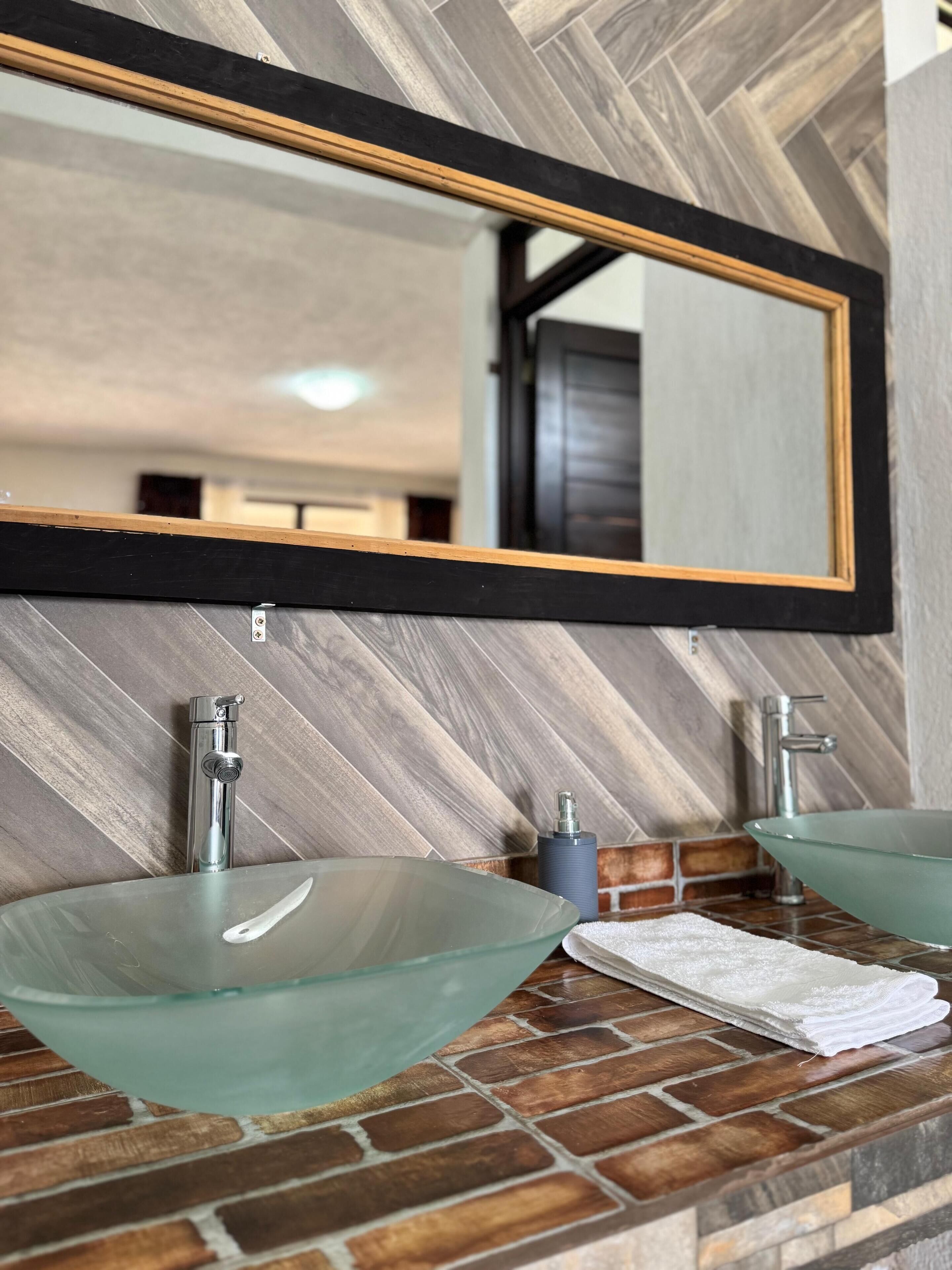 Un baño con dos lavamanos de vidrio, un espejo con marco de madera y una toalla en el mostrador.