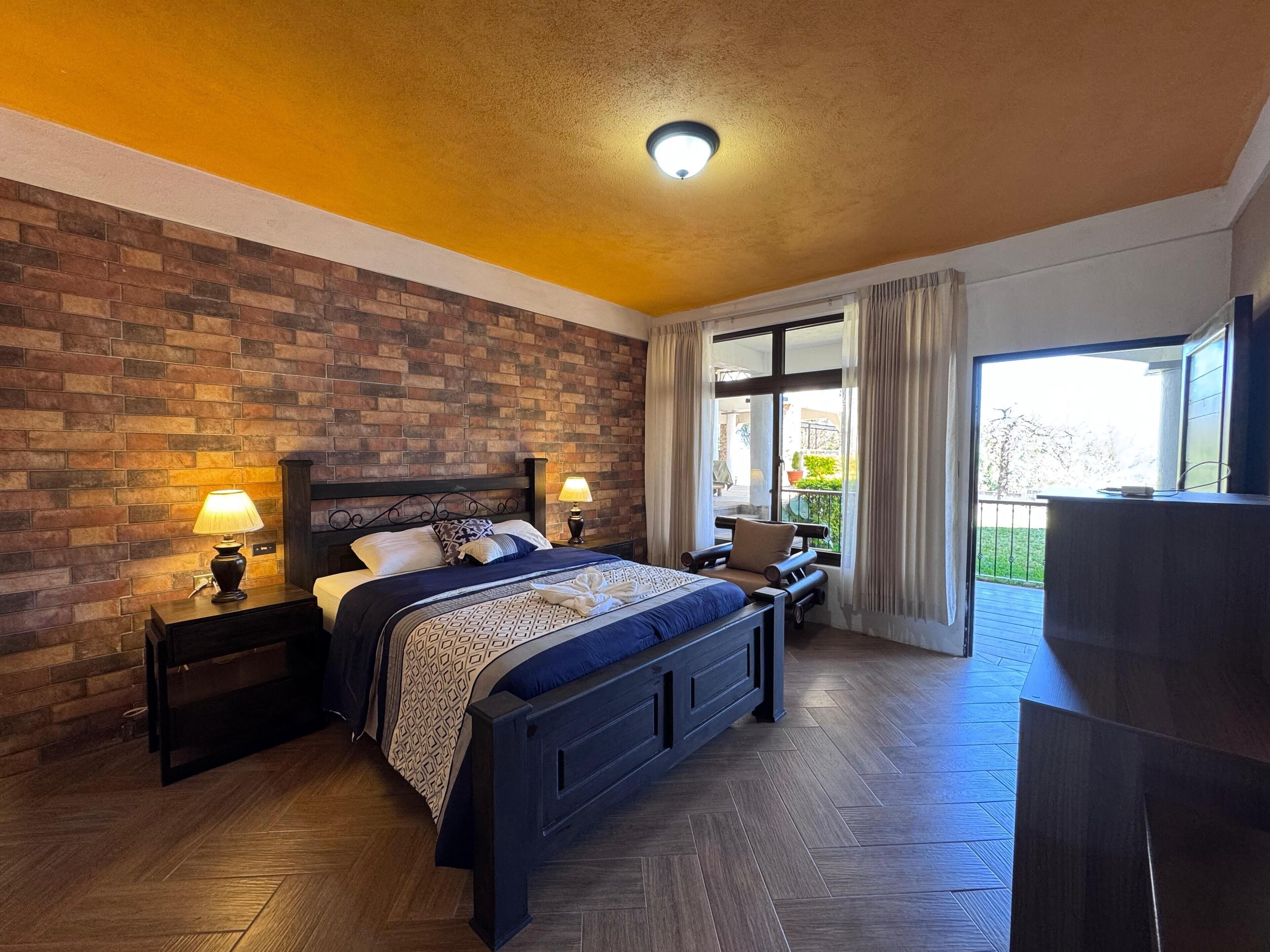Un dormitorio con pared de ladrillo, una cama grande con cabecera, dos mesitas de noche con lámparas, un ventanal con cortinas y un balcón con barandilla.