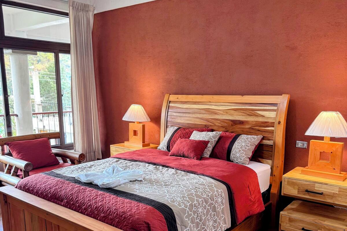 Een houten bed met een rood-wit gebedrukte sprei, twee nachtkastjes met lampen, een houten bankje en een groot raam met uitzicht op groen.