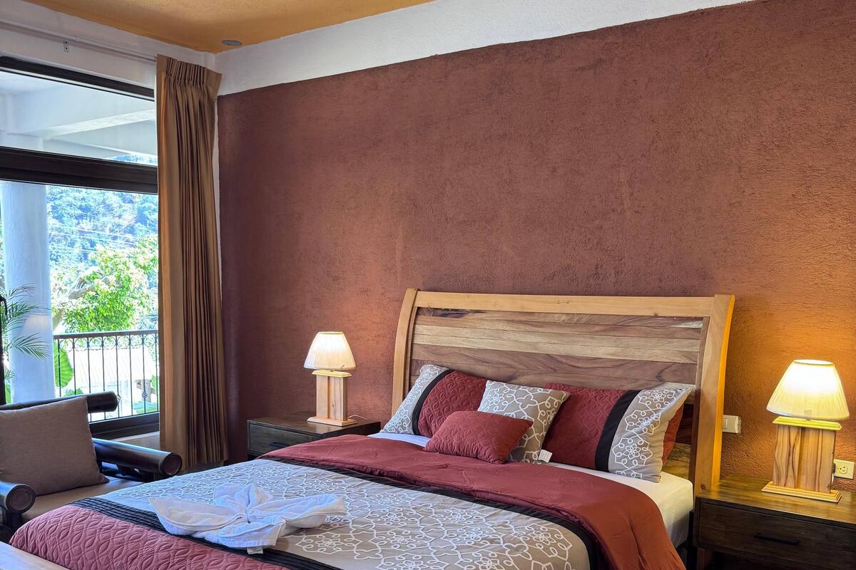 Een slaapkamer met een houten bed, twee nachtkastjes met lampen, een raam met gordijnen en uitzicht op groen.