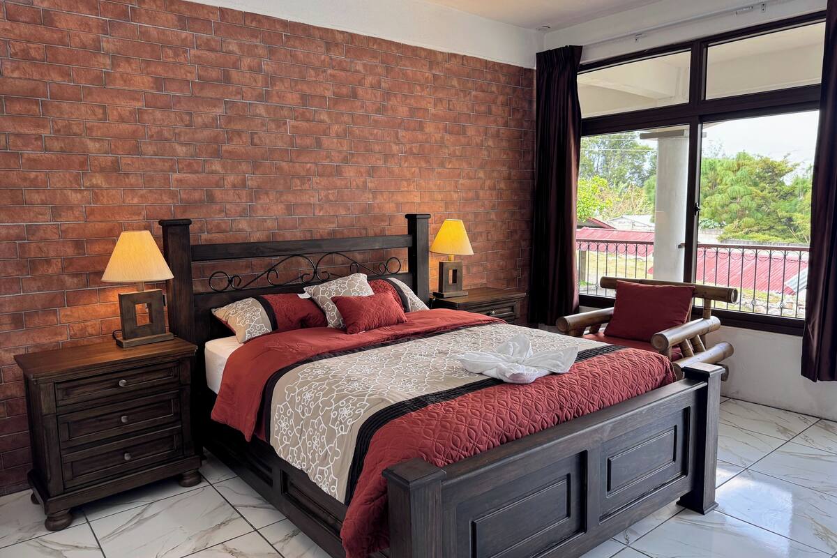 Een slaapkamer met een bakstenen muur, een groot bed met houten hoofdeinde, twee nachtkastjes met lampen en uitzicht op een balkon door een groot raam.