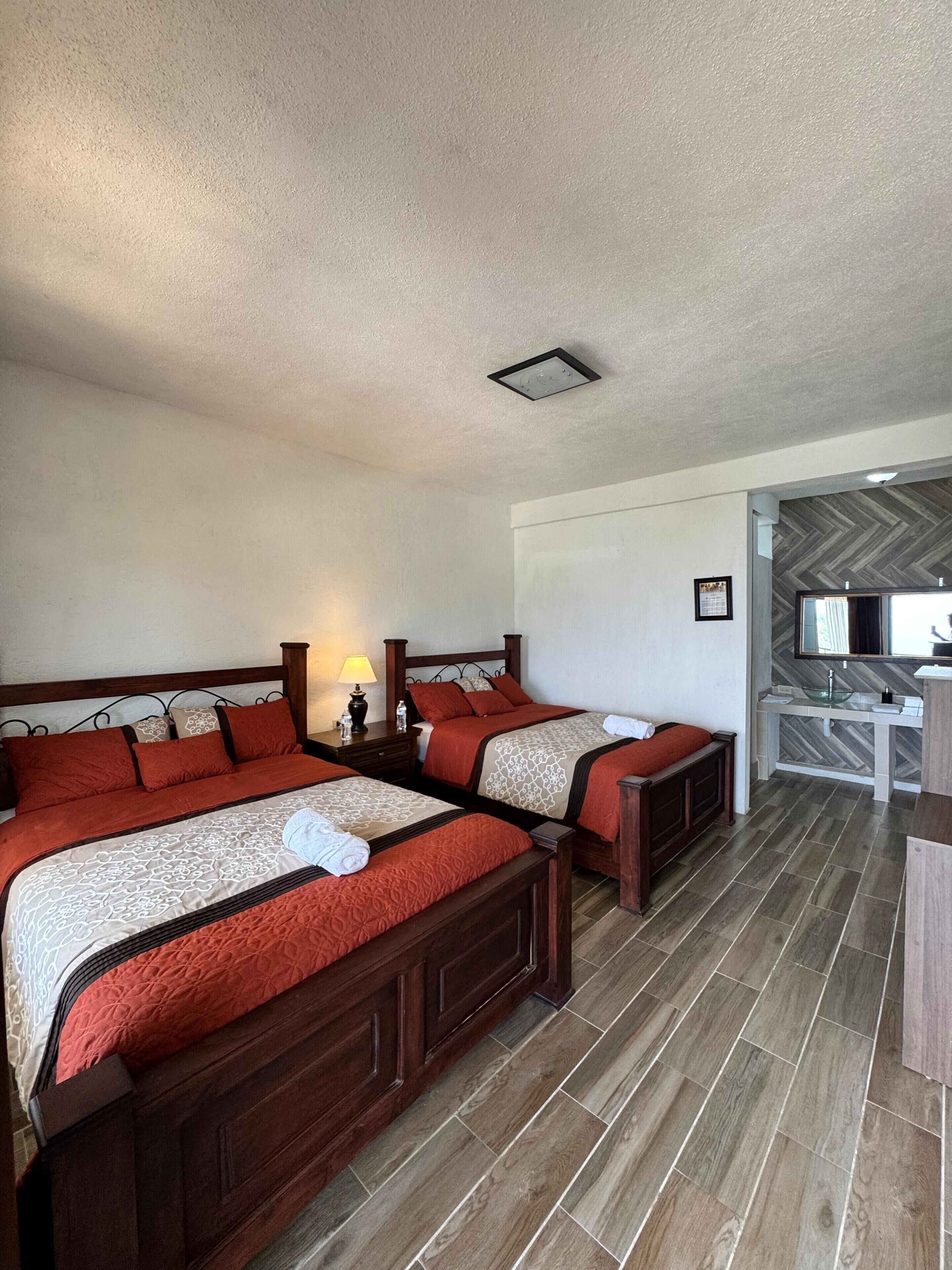 A hotel room with two beds, a wooden floor, a small bathroom with a mirror and sink, and a ceiling light.
