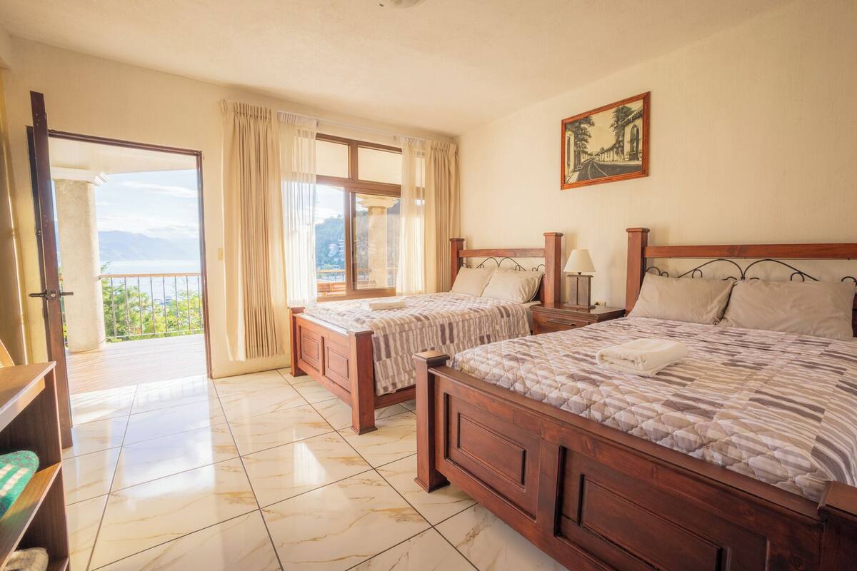 Een hotelkamer met twee houten bedden, een balkon met gordijnen, een schilderij aan de muur en een betegelde vloer.