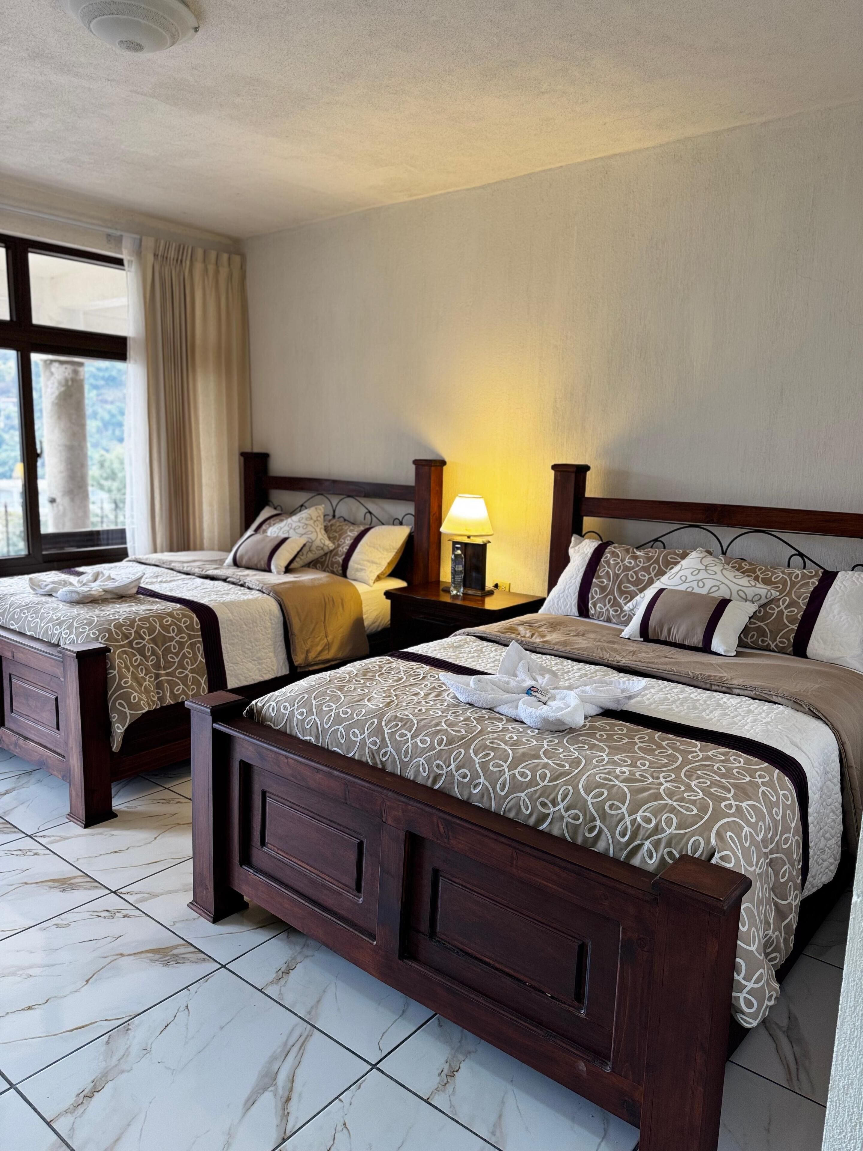 Une chambre d’hôtel avec deux lits, une table de chevet avec une lampe, une fenêtre avec des rideaux et une vue sur l’extérieur.