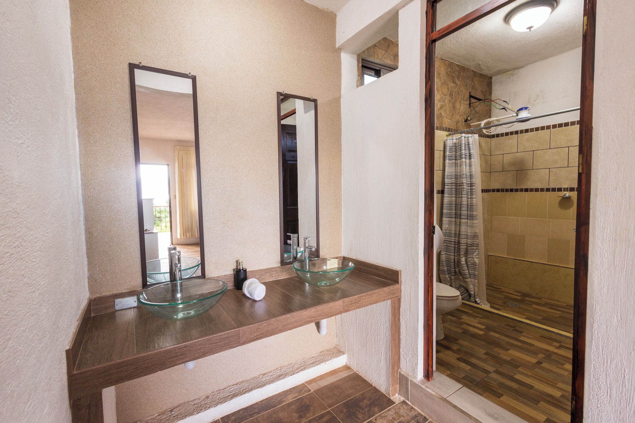 Un baño con un lavabo de cristal, dos espejos, ducha con cortina y suelo de madera.
