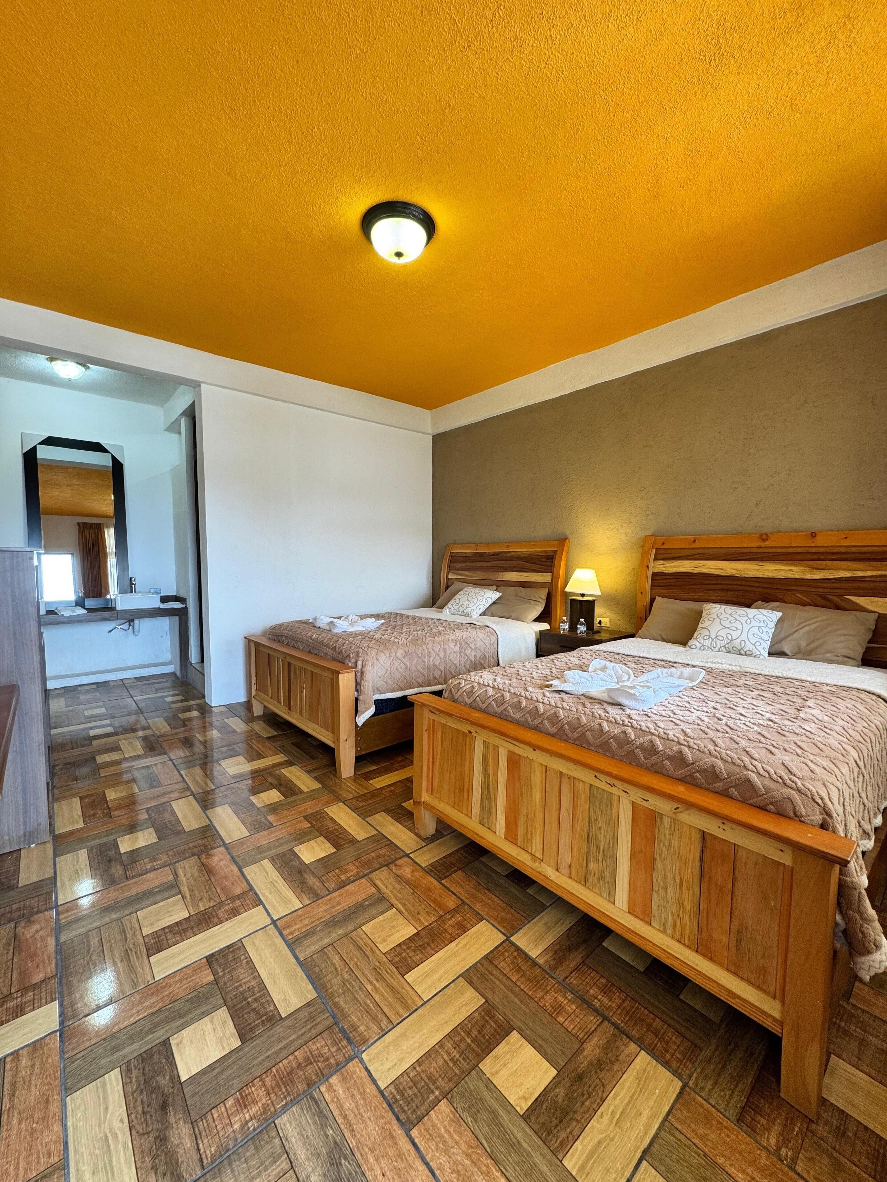 A hotel room with two wooden beds, a herringbone-patterned floor, a ceiling light, and a mirror in the corner.