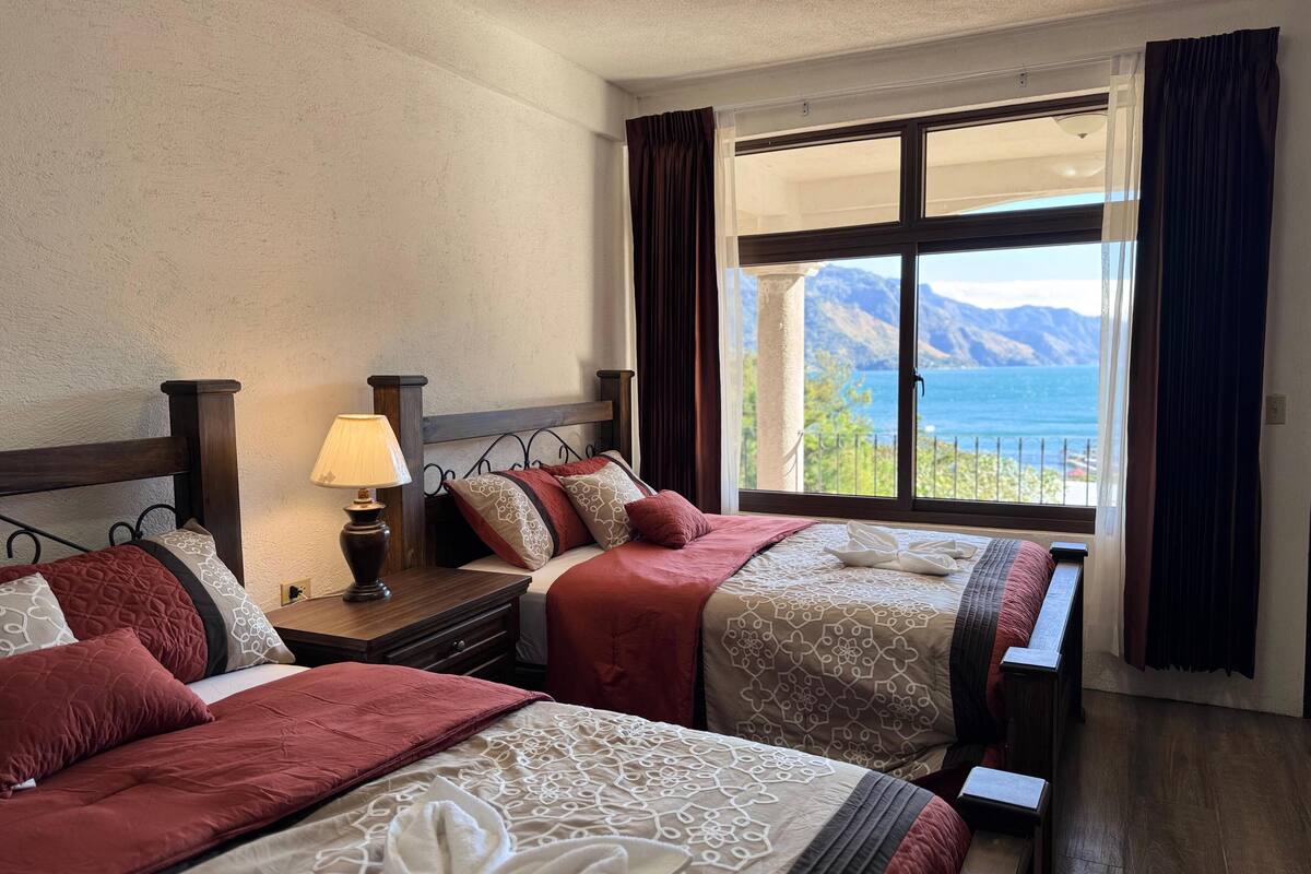 Een hotelkamer met twee bedden, een nachtkastje met een lamp, en een groot raam met uitzicht op zee en bergen.