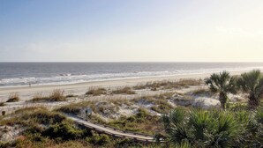 En strand med en träpromenad som leder ner till stranden, omgiven av sanddyner och gles vegetation.