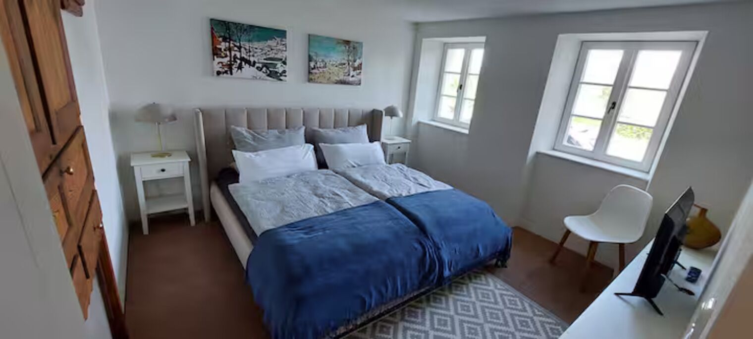Ein Schlafzimmer mit einem Bett, zwei Nachttischen, einem Sessel, einem Schreibtisch mit Computer und zwei Fenstern.