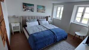 Un dormitorio con una cama, dos mesitas de noche, una silla, un escritorio con una computadora y dos ventanas.