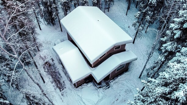 Una cabaña de dos pisos con techo nevado, rodeada por un bosque.