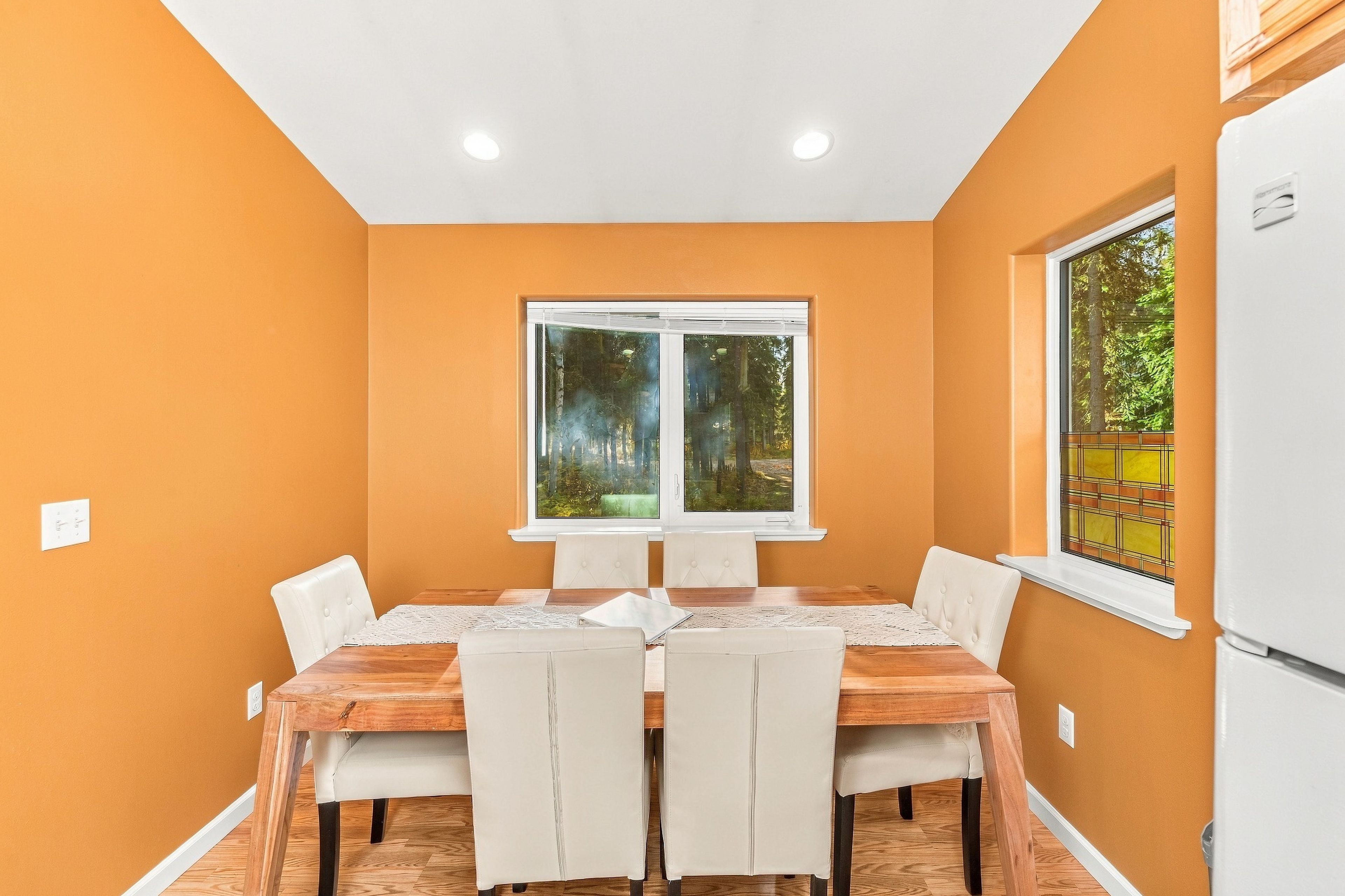 A dining area with a wooden table and chairs, a window with a view of trees, and a white refrigerator.