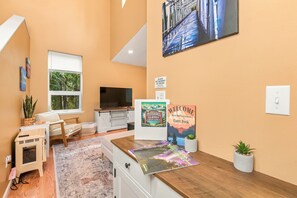 A hotel room with a large window, a flat-screen TV, a wooden desk, a small sofa, a pet carrier, a welcome sign, and a wall-mounted painting.