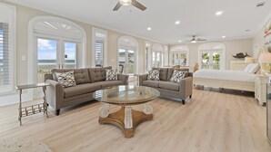 Un salon spacieux avec une grande table basse ronde, deux canapés ornés de coussins à motifs, un ventilateur de plafond et un grand lit en arrière-plan.