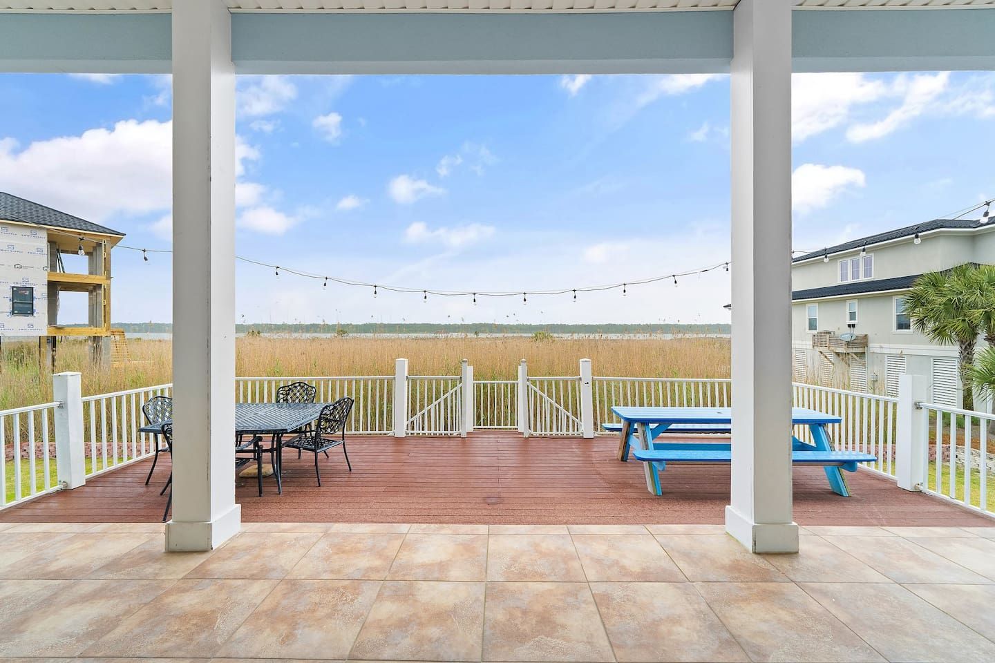 A covered patio with a view of a field, featuring a table and chairs, a blue picnic table, and a white railing.