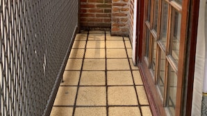 A narrow outdoor corridor with a tiled floor, a metal fence, a brick wall, and a wooden-framed window.