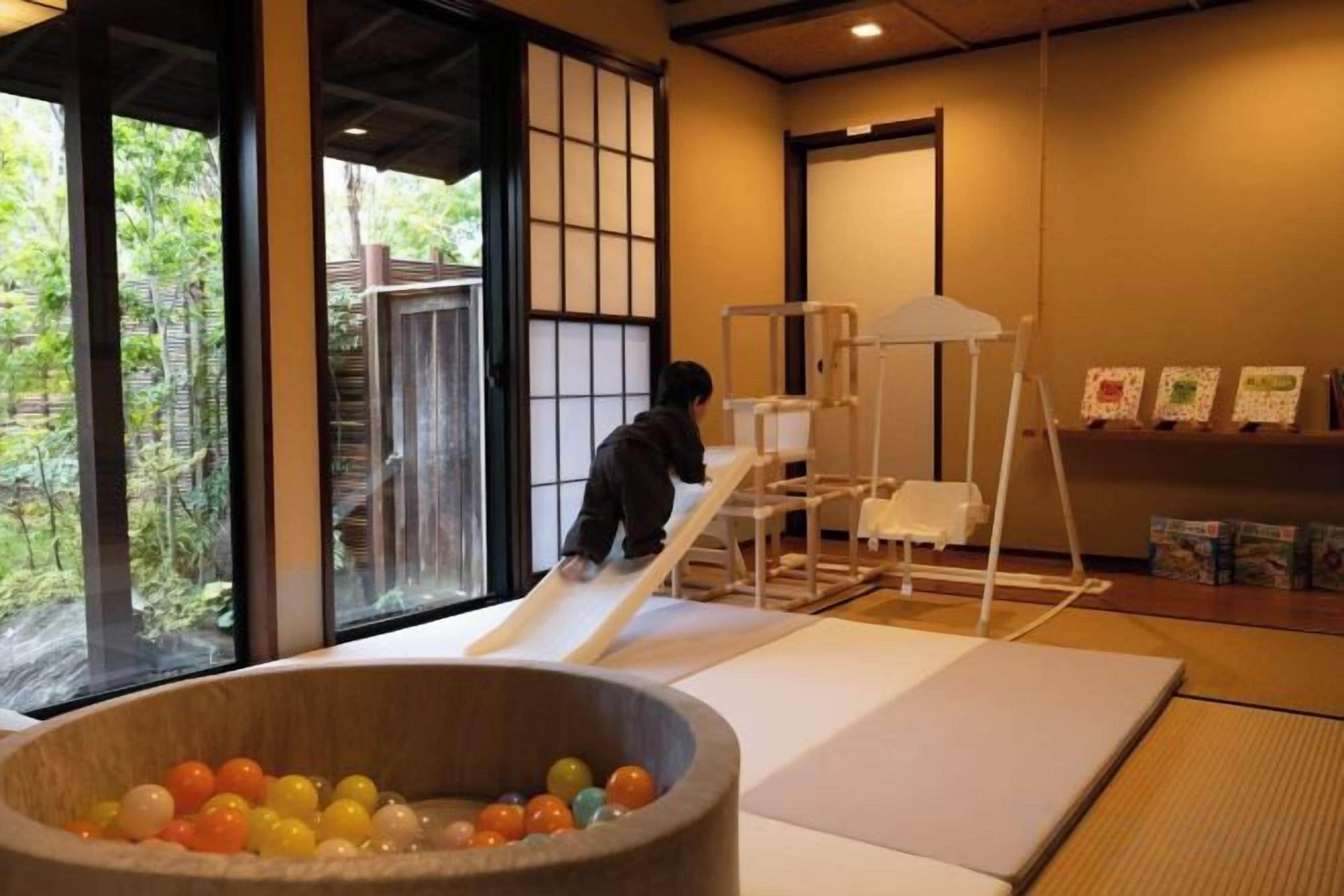 Een kind speelt op een glijbaan in een ruimte met tatami vloerbedekking, een grote houten kom vol met kleurrijke ballen en uitzicht op groen buiten.