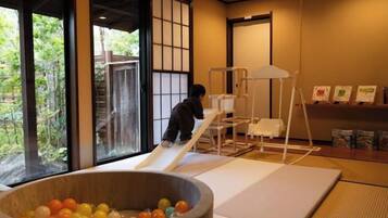 Een kind speelt op een glijbaan in een ruimte met tatami vloerbedekking, een grote houten kom vol met kleurrijke ballen en uitzicht op groen buiten.