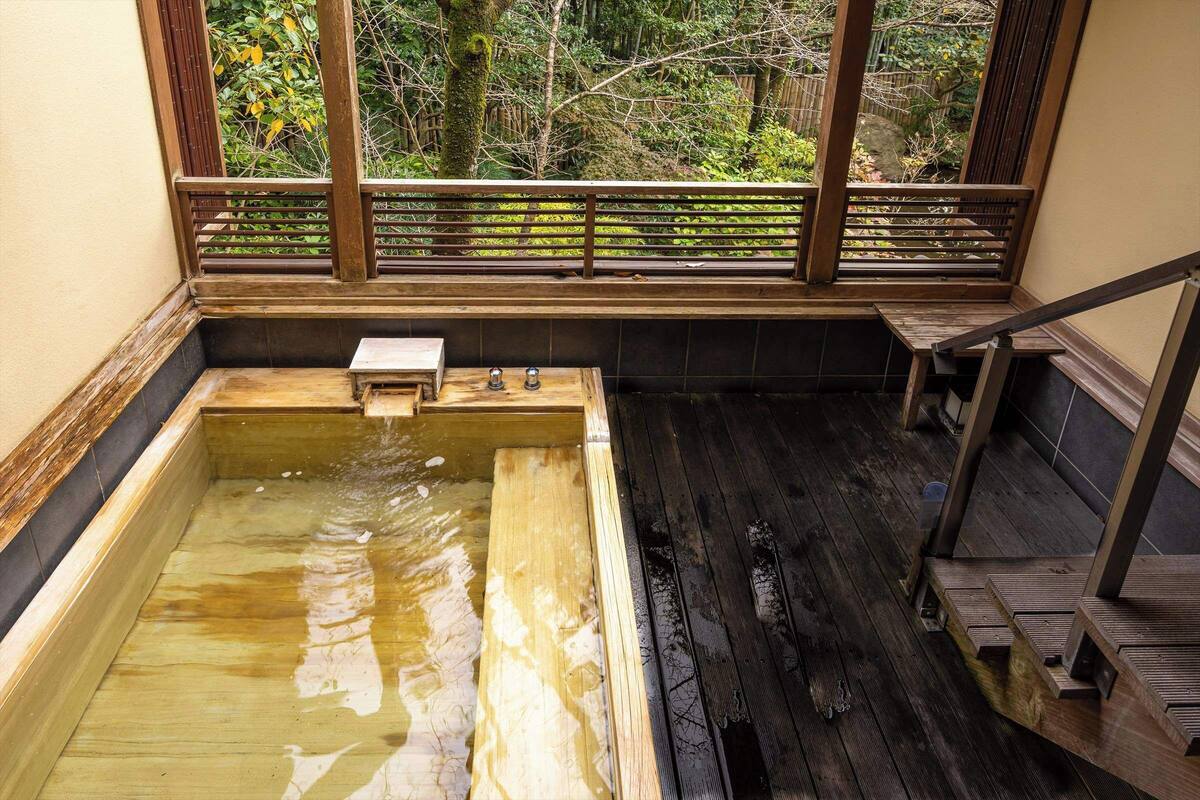 Een houten jacuzzi met een houten bankje en een klein tafeltje, omringd door een houten leuning en uitzicht op bomen door een raam.