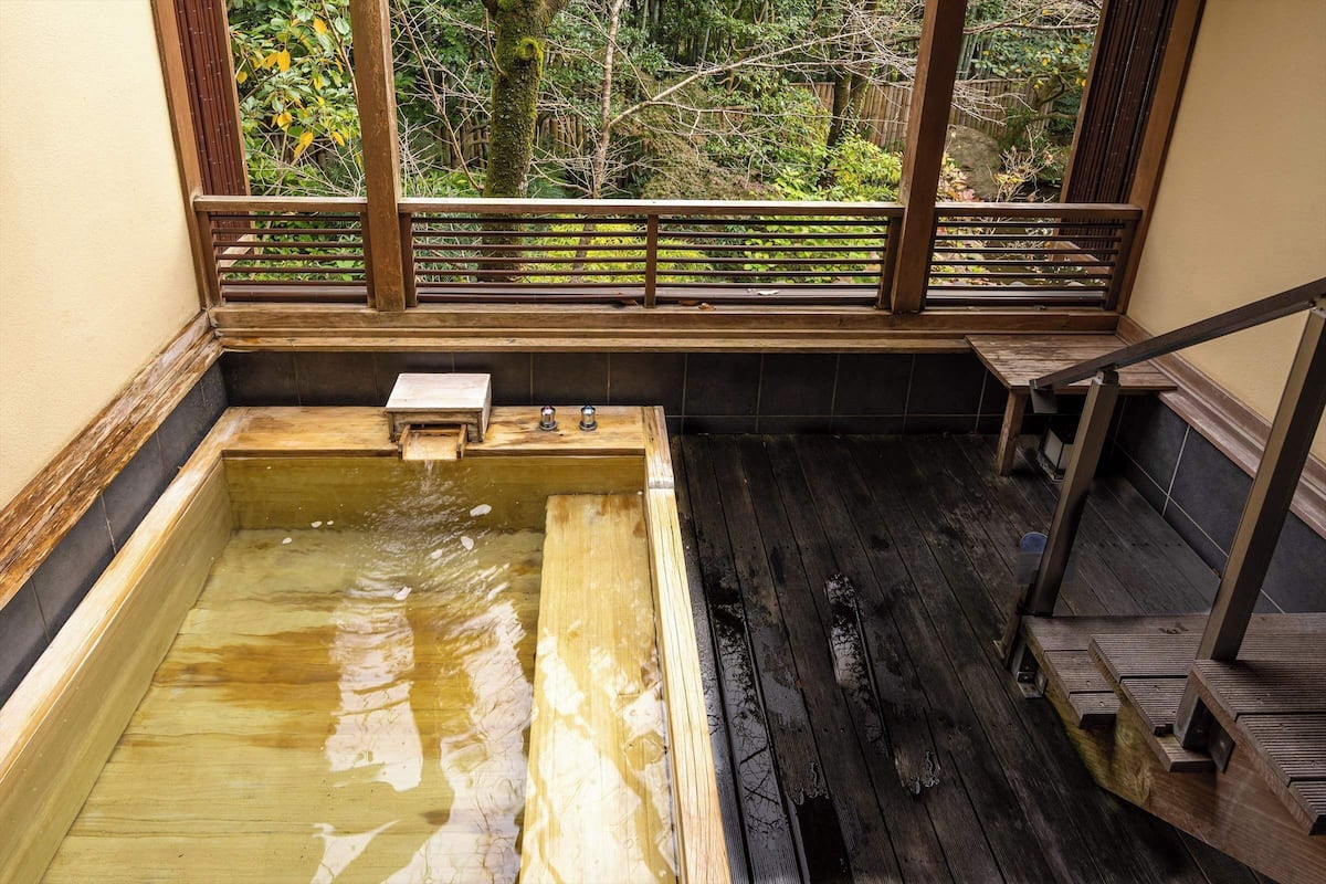 Un bain à remous en bois, doté d’un banc en bois et d’une petite table, entouré d’une balustrade en bois et offrant une vue sur les arbres à travers une fenêtre.