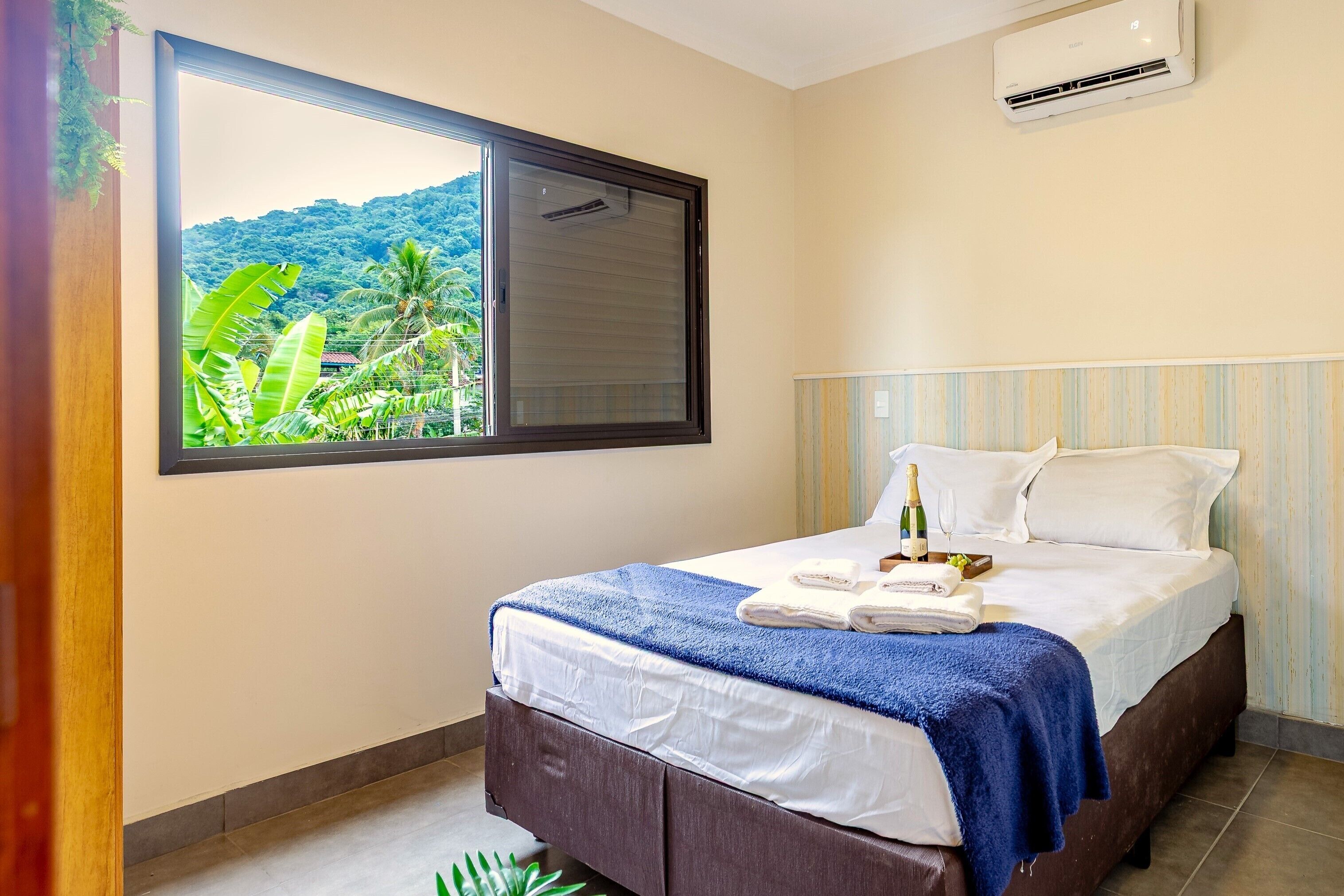Quarto de hotel com uma janela grande, cama com roupa de cama branca, toalha azul e uma garrafa de vinho sobre a mesa de cabeceira.